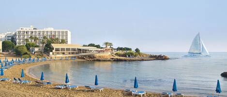 A beach with sun loungers and umbrellas, a sailboat, and a hotel in the background.
