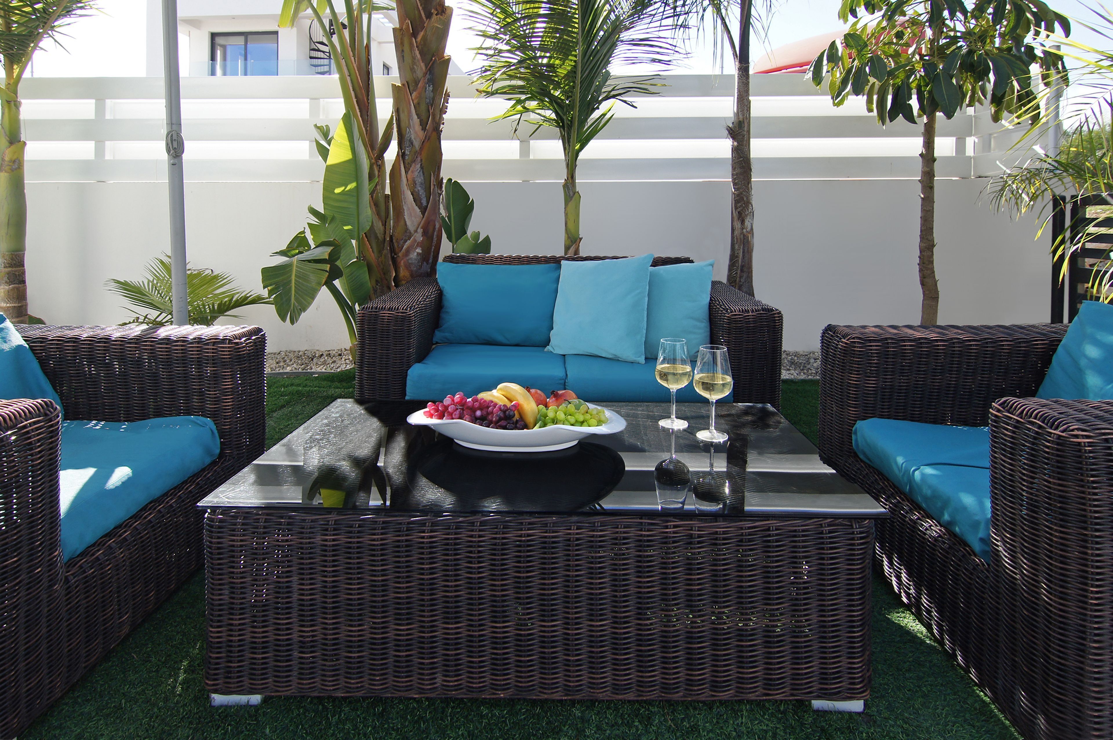 Un espacio de descanso al aire libre con mobiliario de mimbre, un sofá azul, una mesa de centro con fruteros y copas de vino, y plantas tropicales.