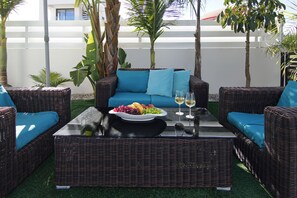Un espace de détente extérieur aménagé avec des meubles en osier, comprenant un canapé bleu, une table basse avec un panier de fruits et des verres à vin, ainsi que des plantes tropicales.