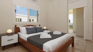 A neatly made bed with a wooden headboard, a nightstand with a lamp, a window with a view of buildings, and a door leading to a balcony. - VILLA CLEOPATRA  - 4bed Villa with large pool in Ayia Napa Hills (Paralimni)
