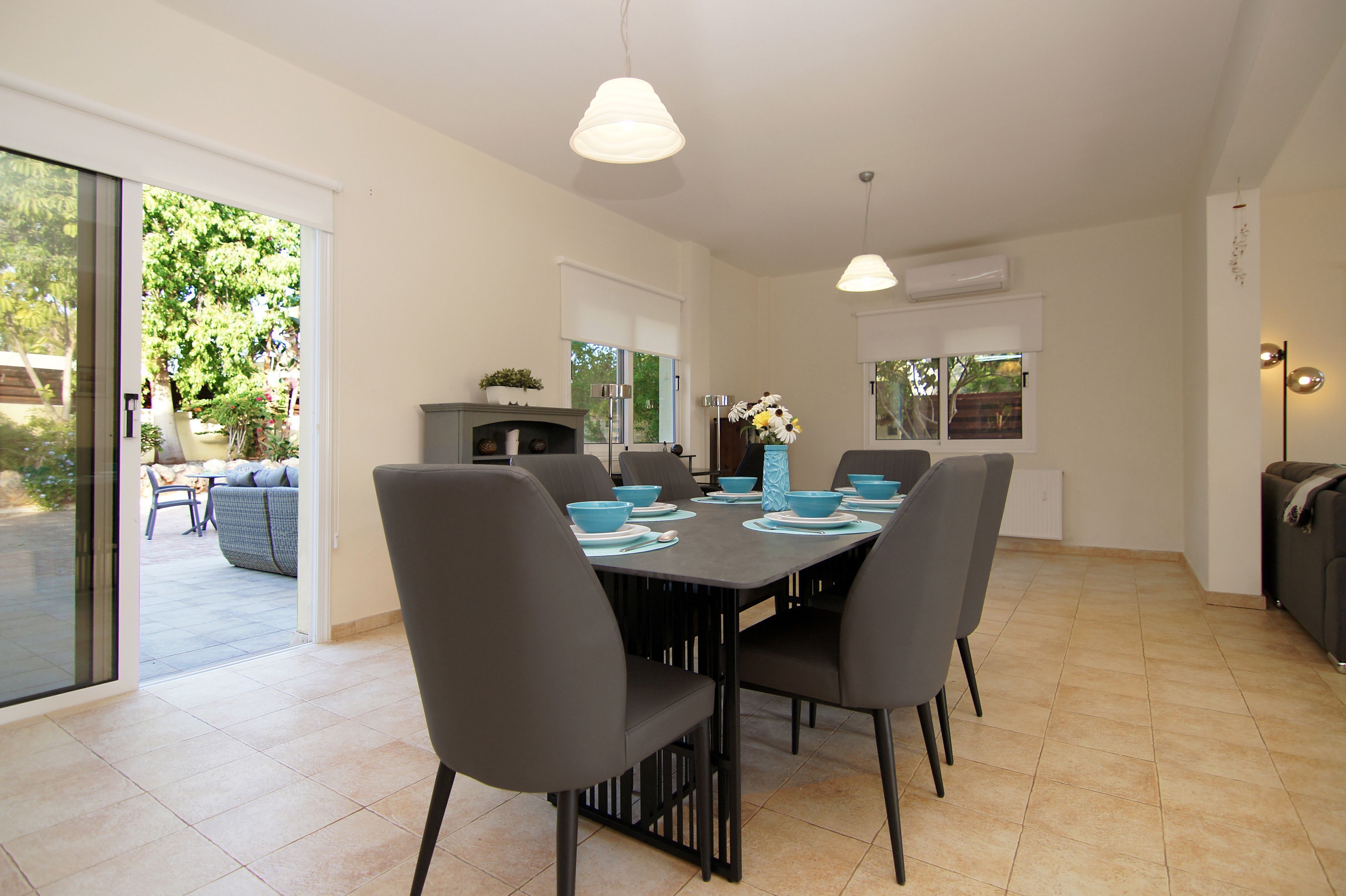 A modern dining area with a large table, grey chairs, and blue bowls. There is a sliding glass door leading to an outdoor patio with a sofa and a dining table.
