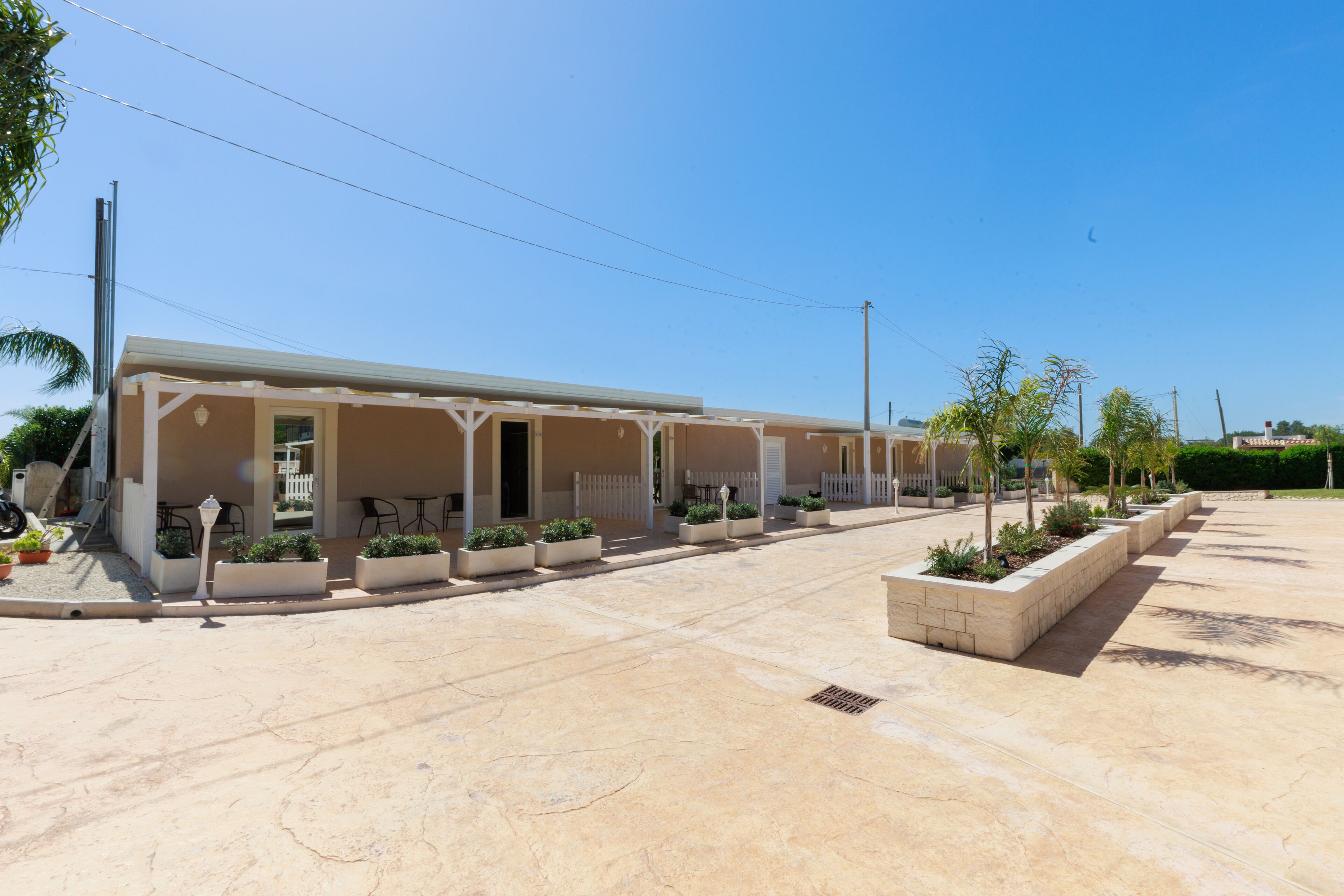 A row of single-story buildings with outdoor seating areas, a paved courtyard, and planters with small trees.