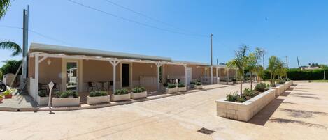 A row of single-story buildings with outdoor seating areas, a paved courtyard, and planters with small trees.