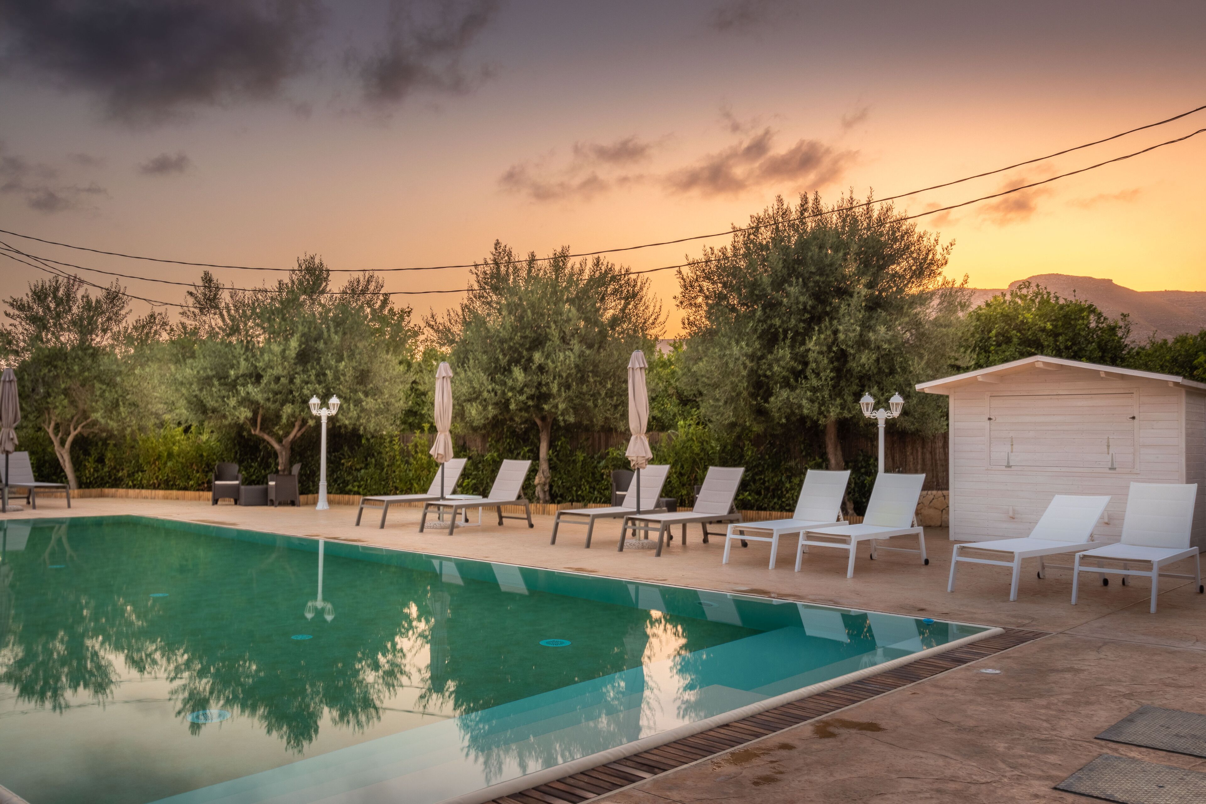 Una piscina con sillas de descanso y sombrillas, rodeada de árboles y un cobertizo de madera.