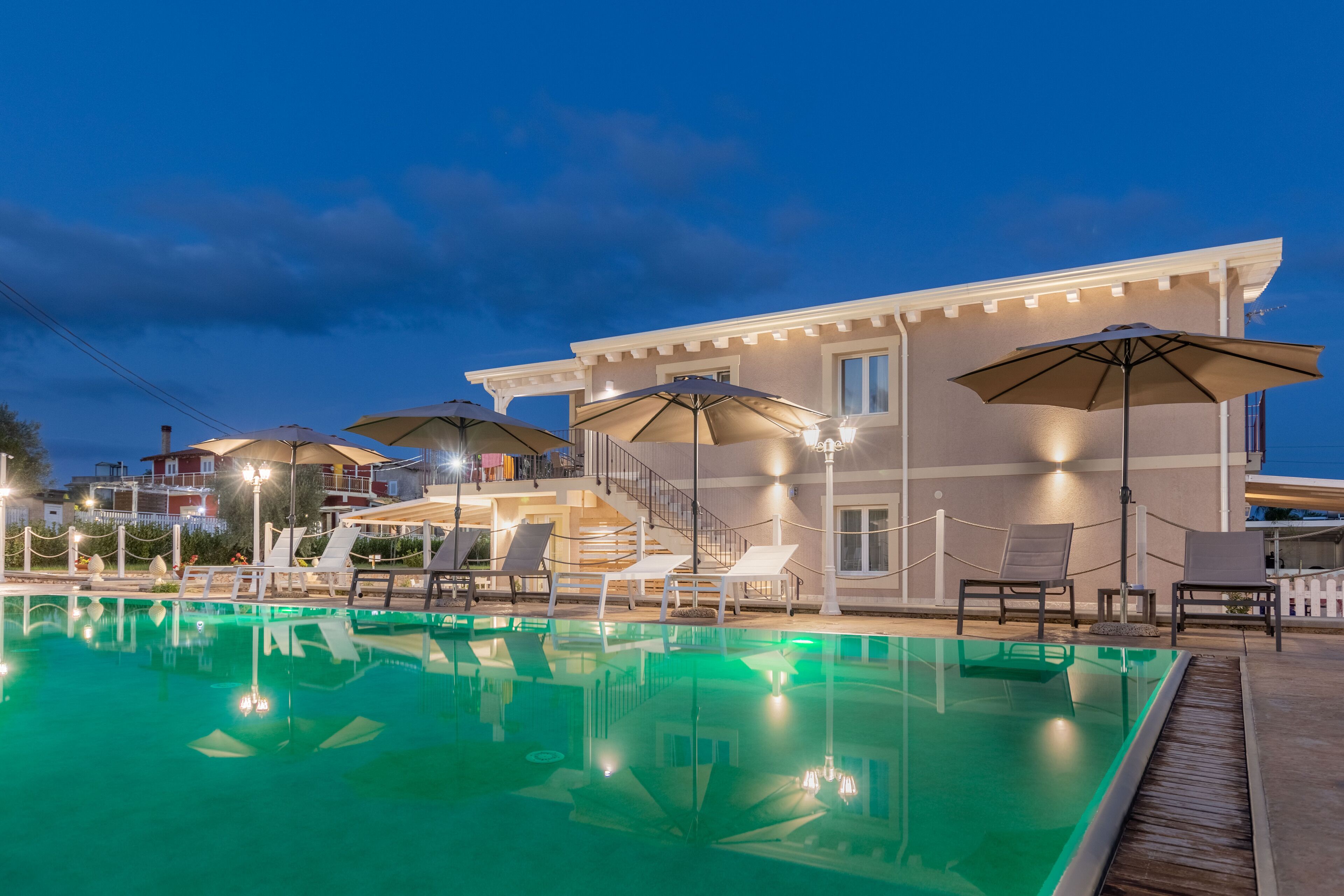 A swimming pool with lounge chairs and umbrellas, a covered seating area, and a building with a balcony.