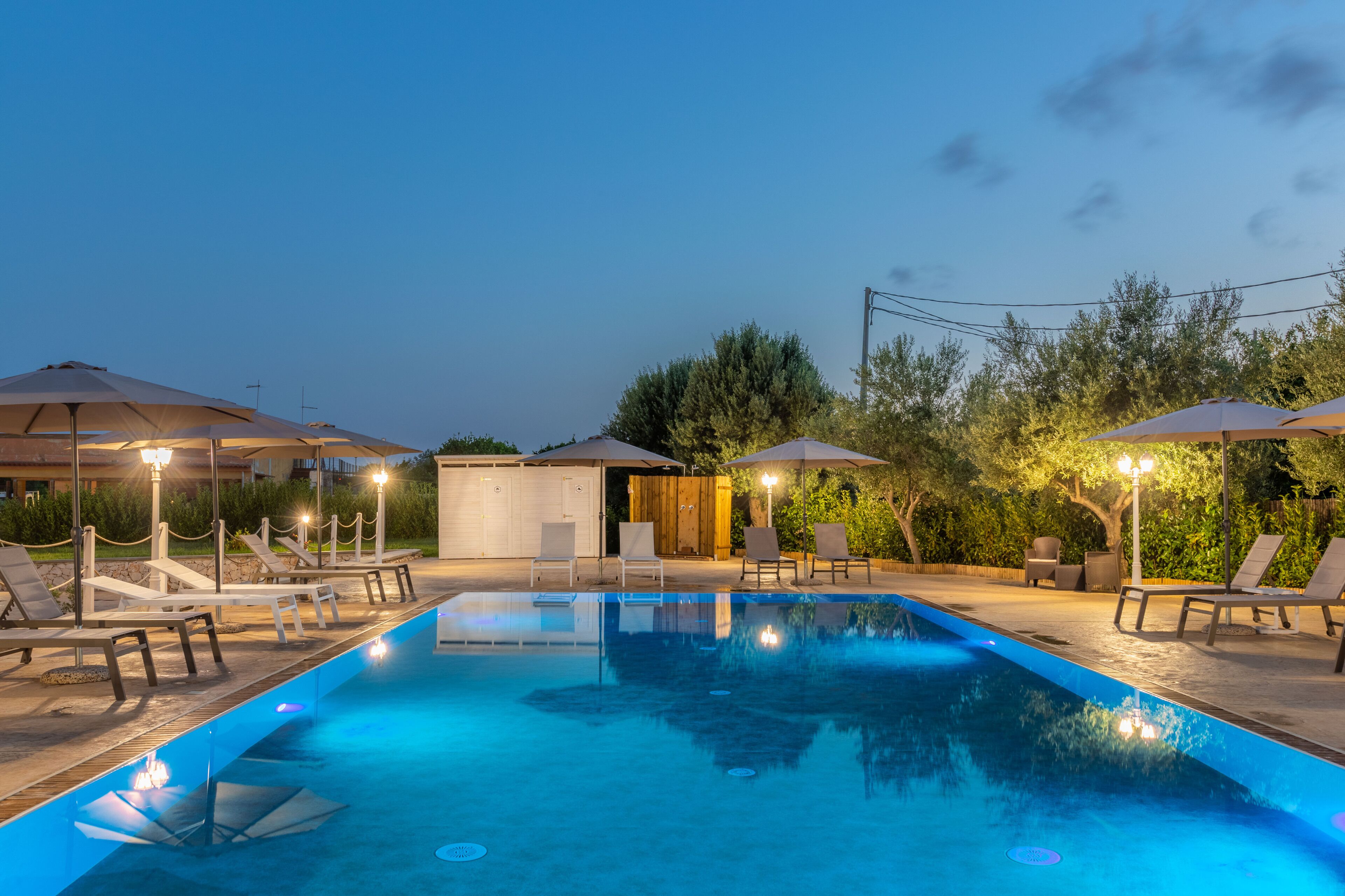 A swimming pool with lounge chairs and umbrellas, surrounded by greenery and a wooden building.