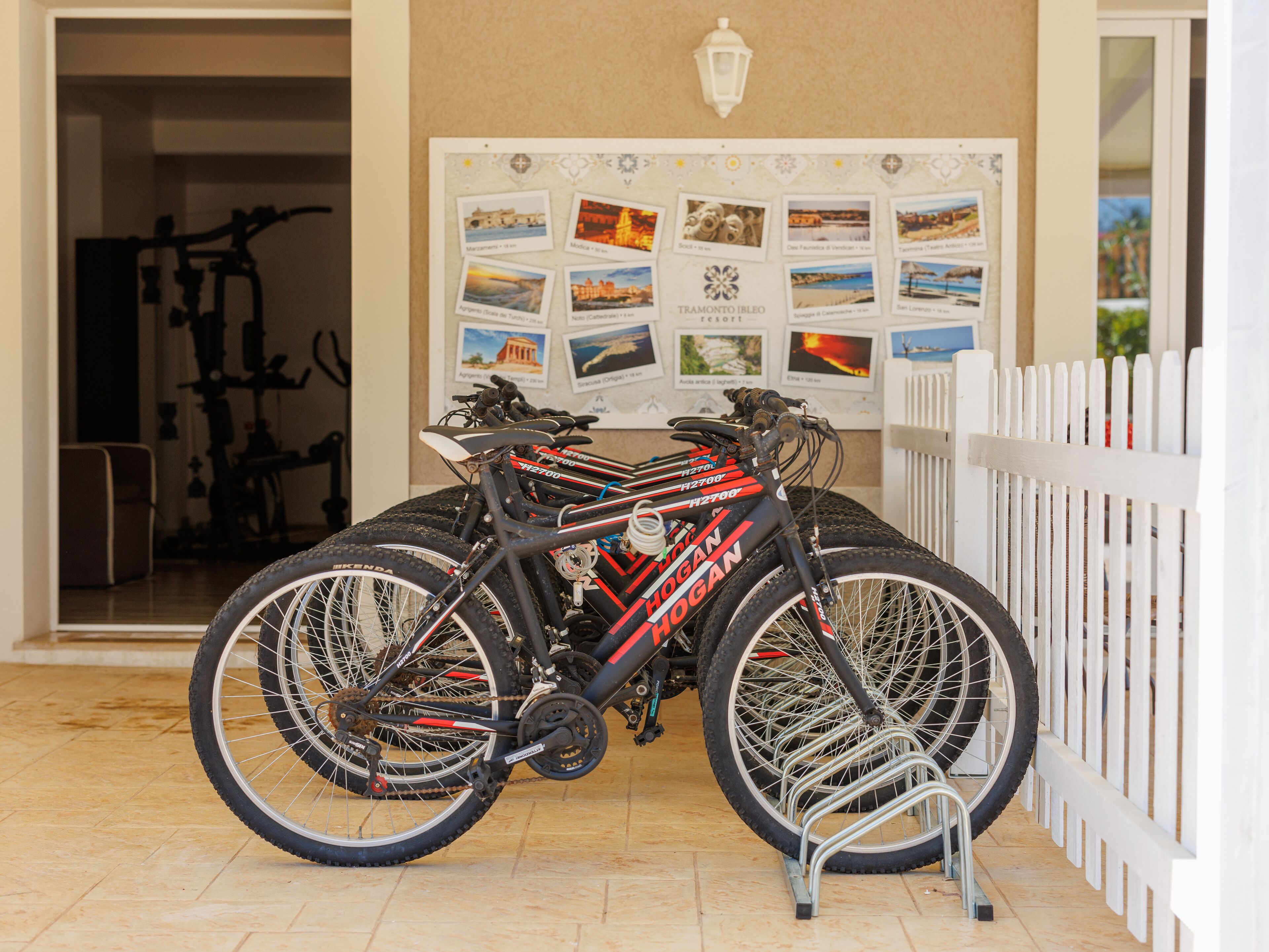 Une pièce avec un porte-vélos où sont rangés deux vélos, un tableau d’affichage avec des photos et une clôture blanche.