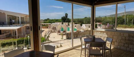 A covered outdoor area with tables and chairs, large windows offering views of a pool and greenery.