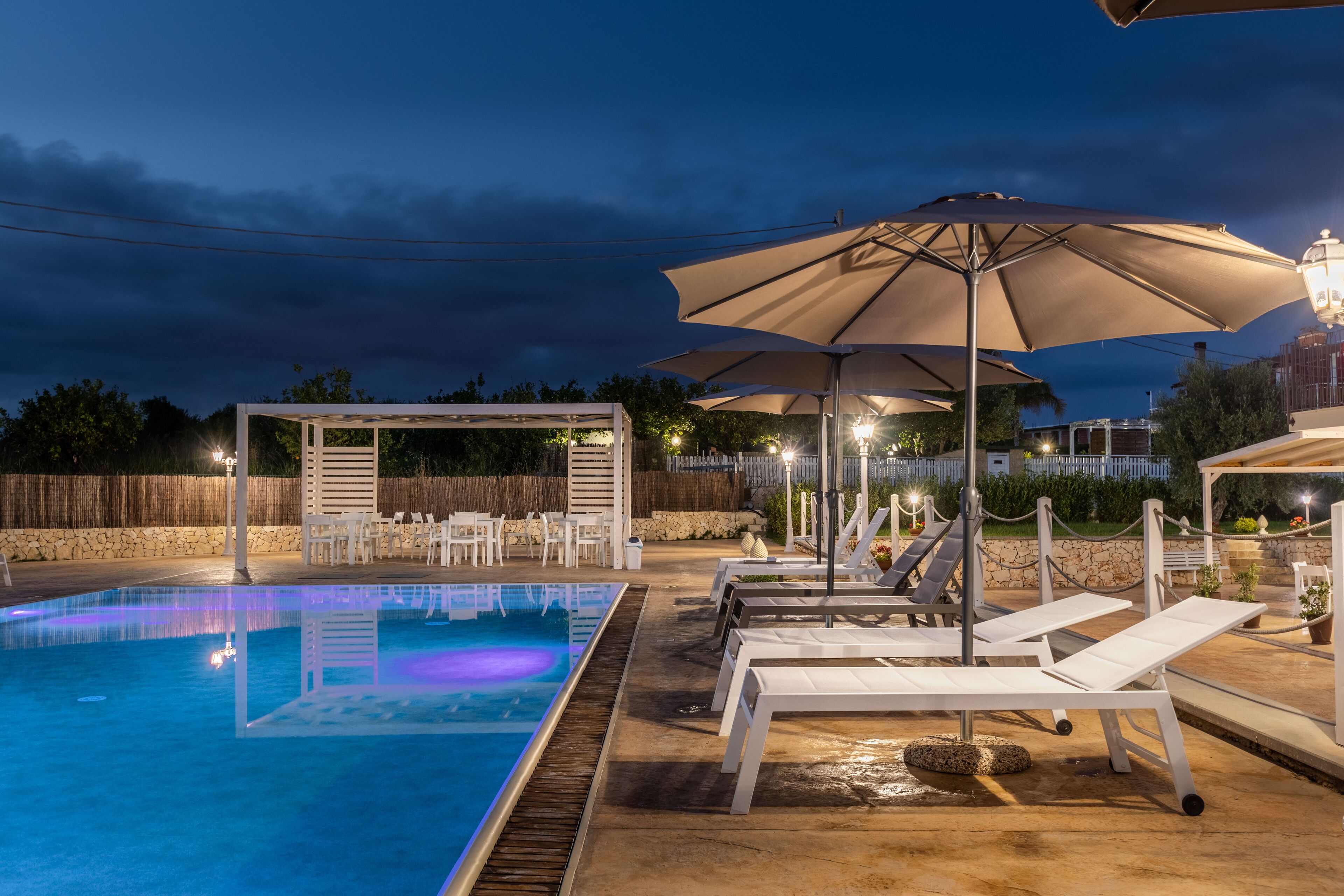 A swimming pool with lounge chairs and umbrellas, surrounded by a stone wall and wooden fence.