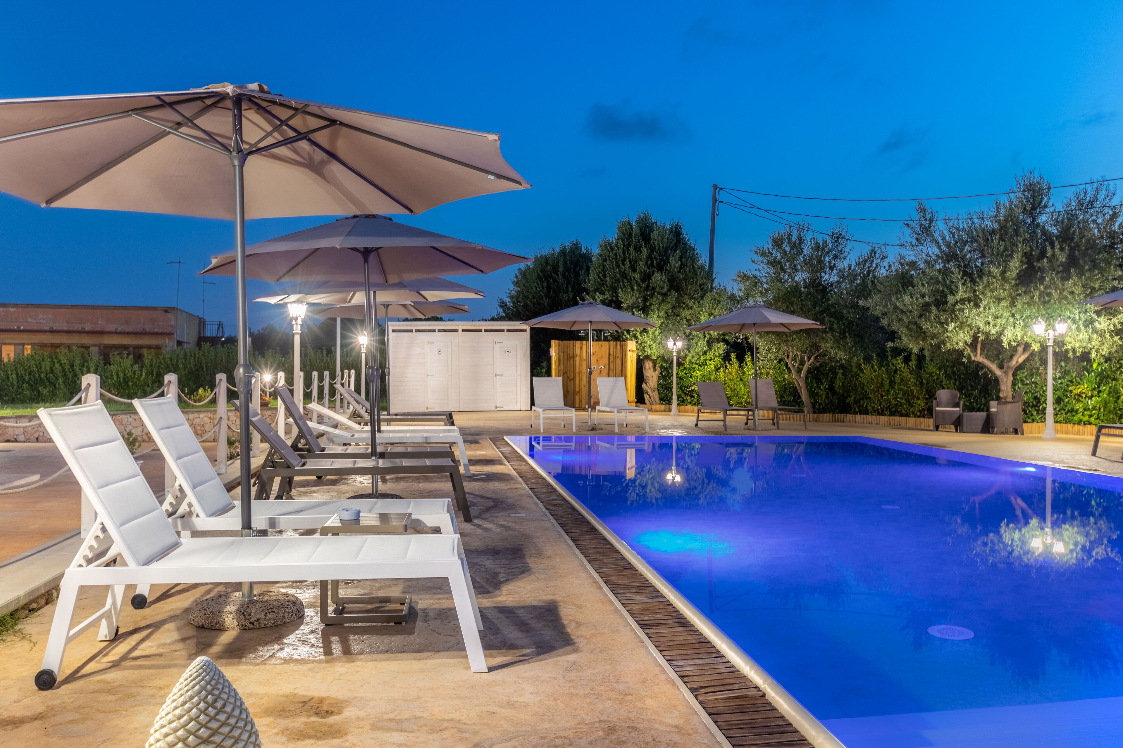 A swimming pool area with lounge chairs and umbrellas, surrounded by greenery and trees.