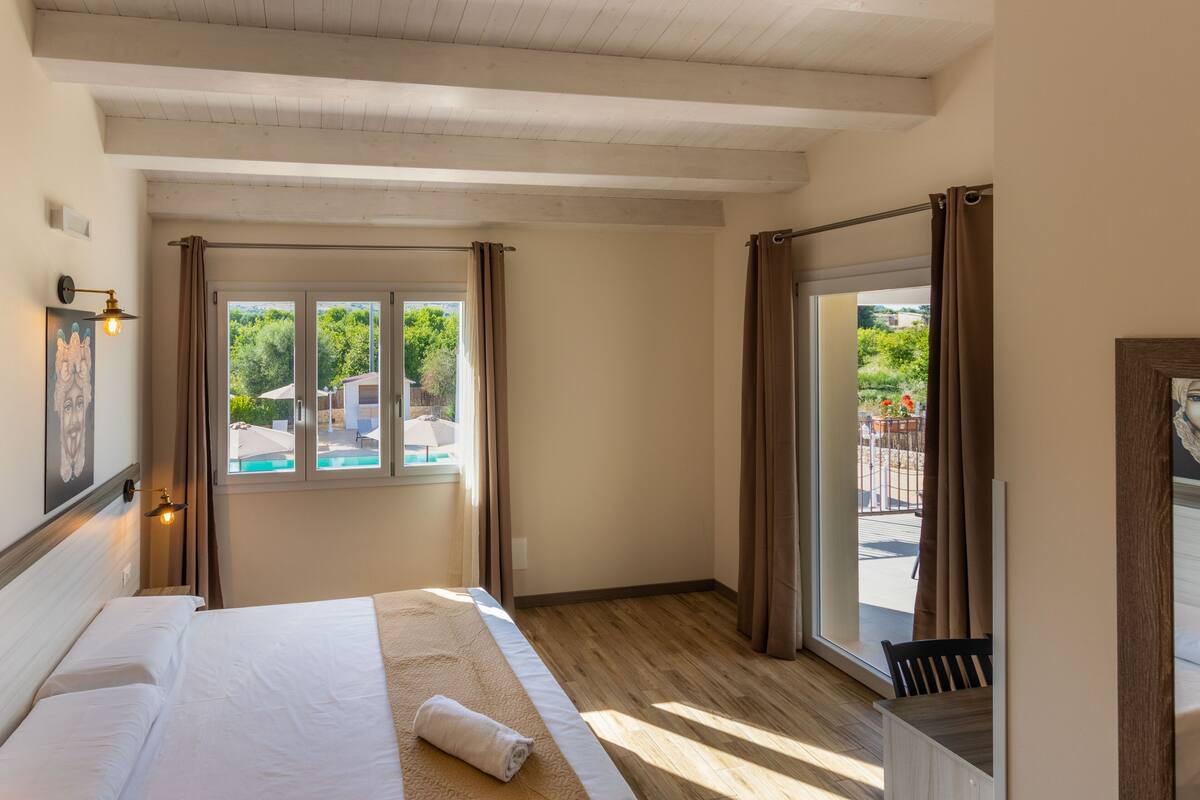 A hotel room with a bed, a window with curtains, a wooden floor, and a view of a swimming pool and greenery outside.