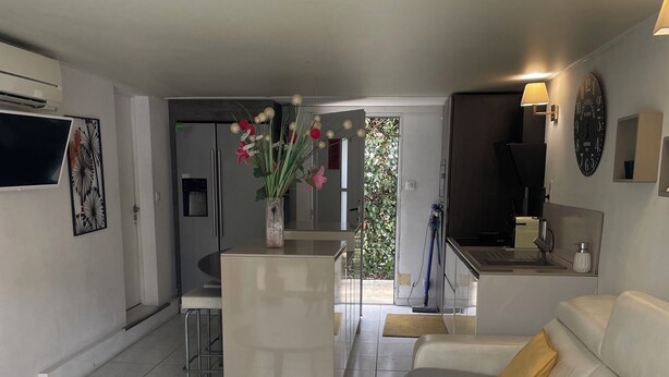Een moderne woonkamer met een salontafel met glazen blad, een bank met kussens, een eethoek met een tafel en stoelen, een keuken met een spoelbak en fornuis, en een grote wandklok.