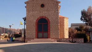 A small stone building with a red arch entrance, a cross on top, and a flag on the left. - House near the bay of Cornino (Cornino)