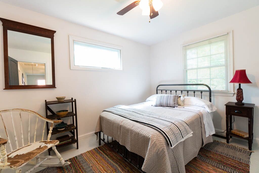 Un dormitorio con cama, una silla, estante, lámpara y ventana.