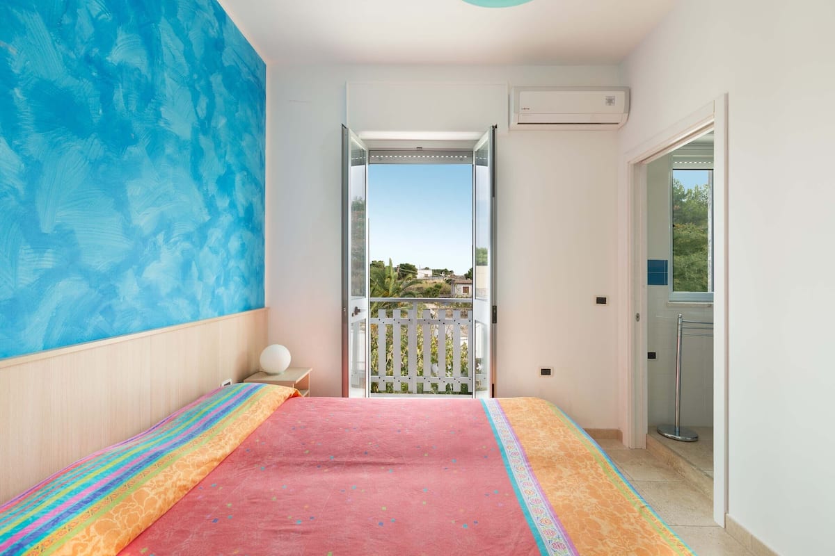 A bedroom with a large bed, a view of the outdoors through an open door, and a balcony with a white railing.