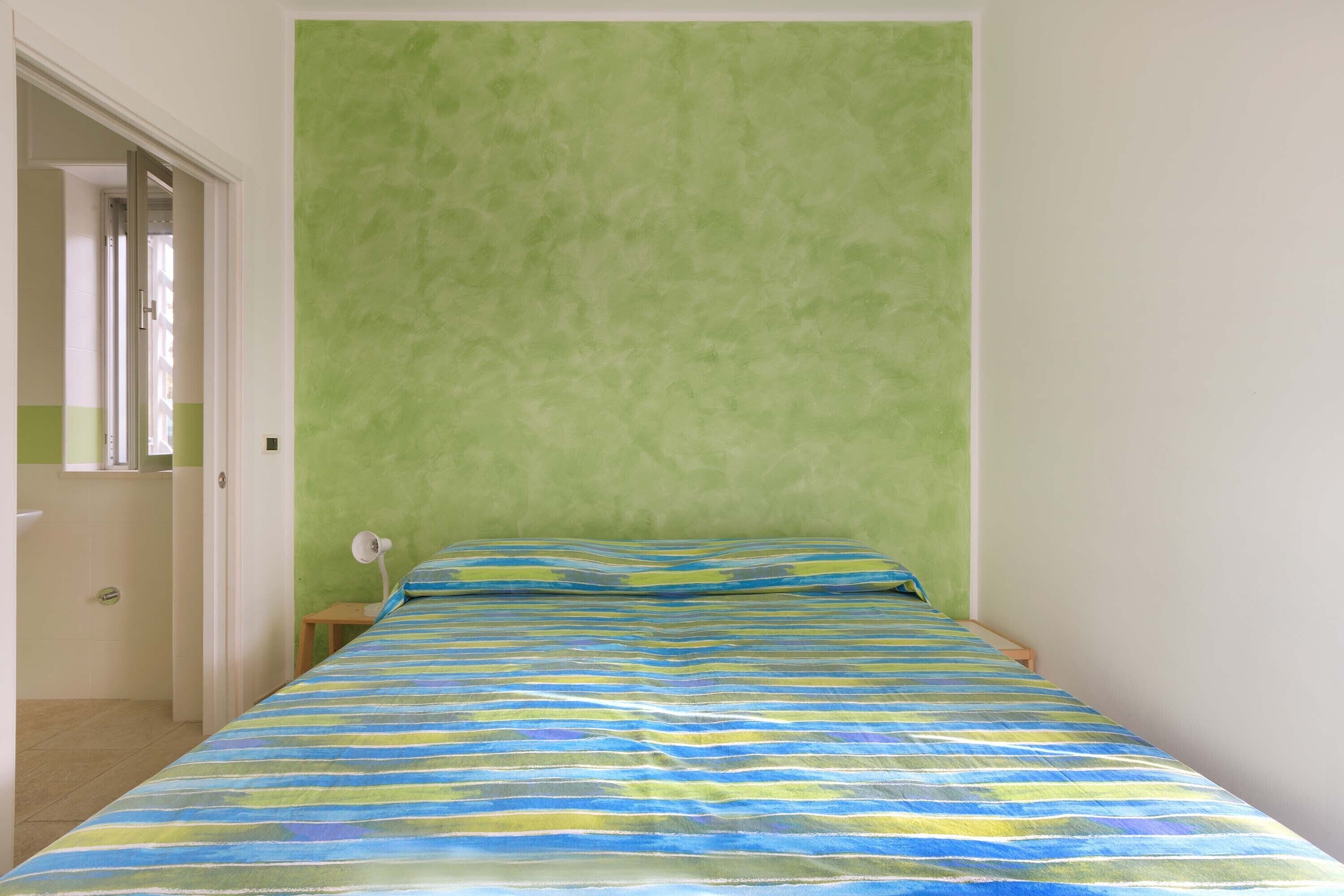 Ein kleines Zimmer mit einem Einzelbett, grüner Wand und einer gestreiften Bettdecke.