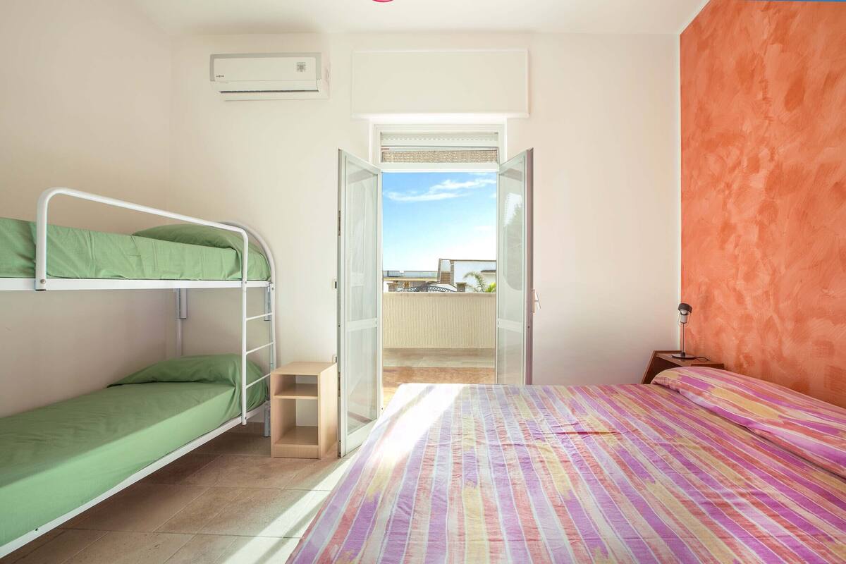 A room with a bunk bed, a single bed, and a window leading to a balcony.