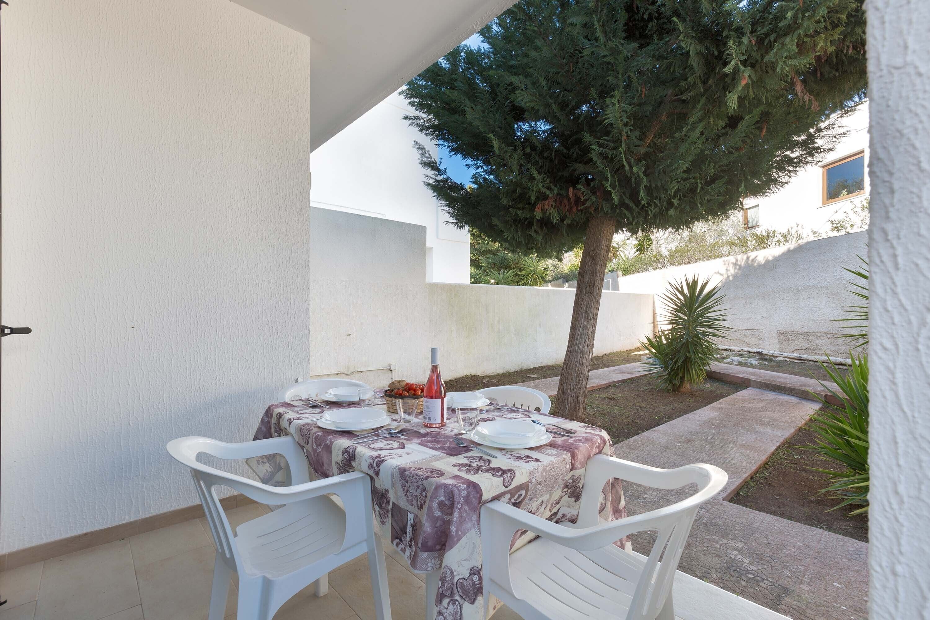 Un patio con una mesa dispuesta para dos personas, rodeado de sillas de plástico blanco, una botella de vino y una cesta de fruta.