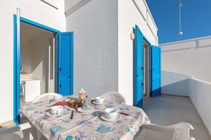 A sunlit outdoor dining area with a table set for two, featuring a white tablecloth with blue patterns, a coffee pot, cups, and a plate of food.