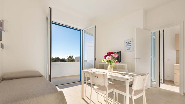 Een moderne kamer met een bed, een eettafel met stoelen, een vaas met bloemen en uitzicht door een glazen deur.