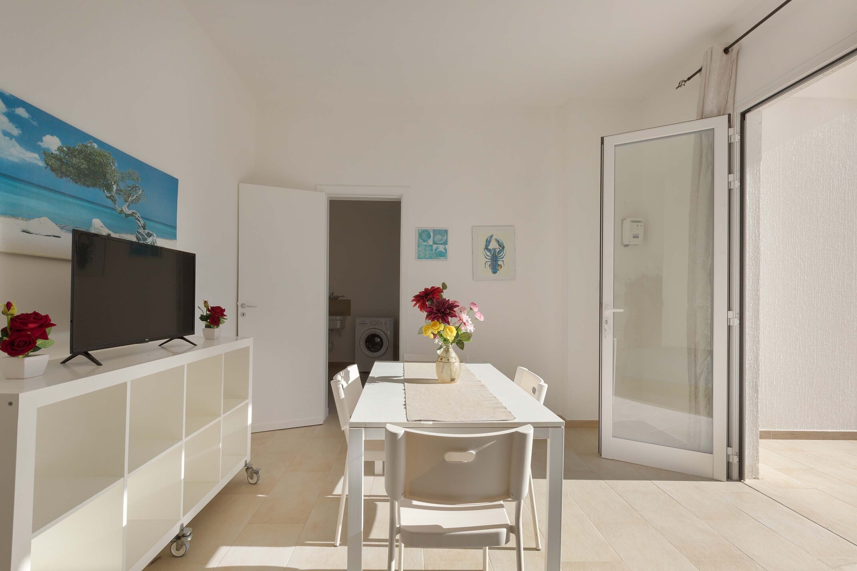 A modern dining area with a white table, chairs, a TV on a white cabinet, and a vase with flowers.