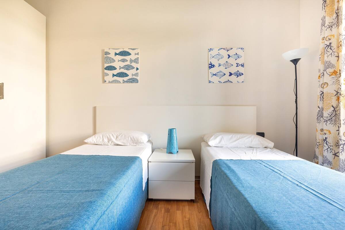 Een kamer met twee eenpersoonsbedden, een witte nachtkastje met een blauwe vaas en wanddecoratie met vissen.