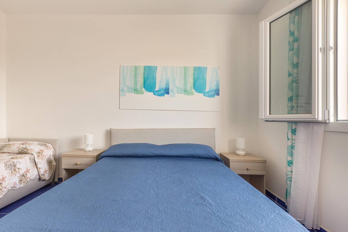 A bedroom with a bed, two bedside tables, a nightstand, a wall-mounted artwork, and a window with a curtain.