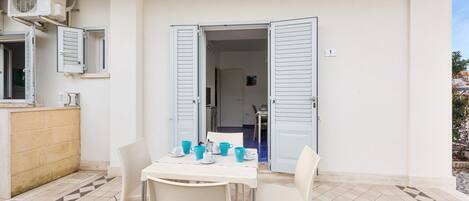 Un pequeño espacio exterior para comer con una mesa y sillas, piso de baldosas y la entrada de un edificio con contraventanas abiertas.
