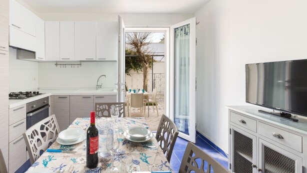 A modern kitchen with white cabinets, a dining table set with plates, a bottle of wine, and a TV on a cabinet. - Residence Spiaggia d'Oro (Porto Cesareo)
