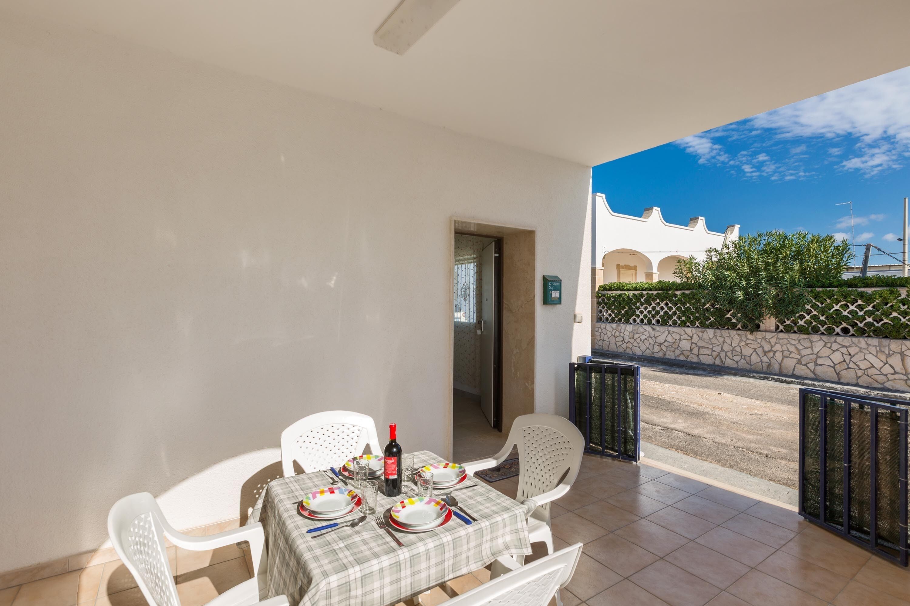A balcony with a table set for dining, white chairs, and a bottle of wine.