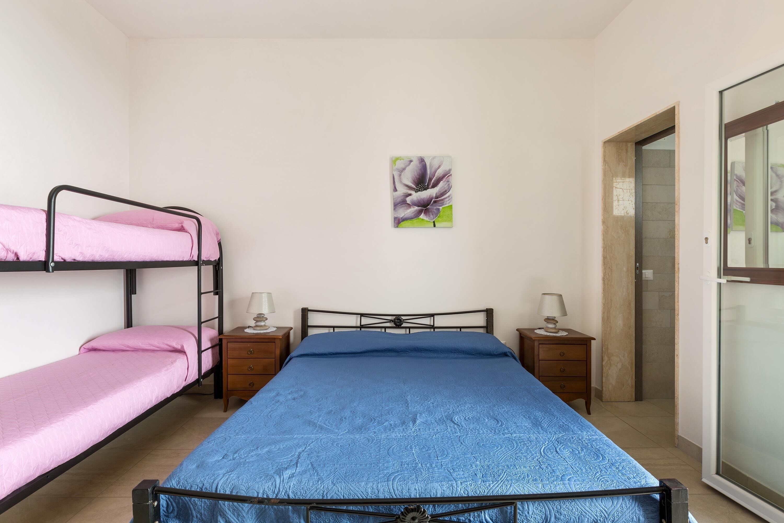 A dormitory room with bunk beds, a single bed, two bedside tables with lamps, and a door leading to a bathroom.