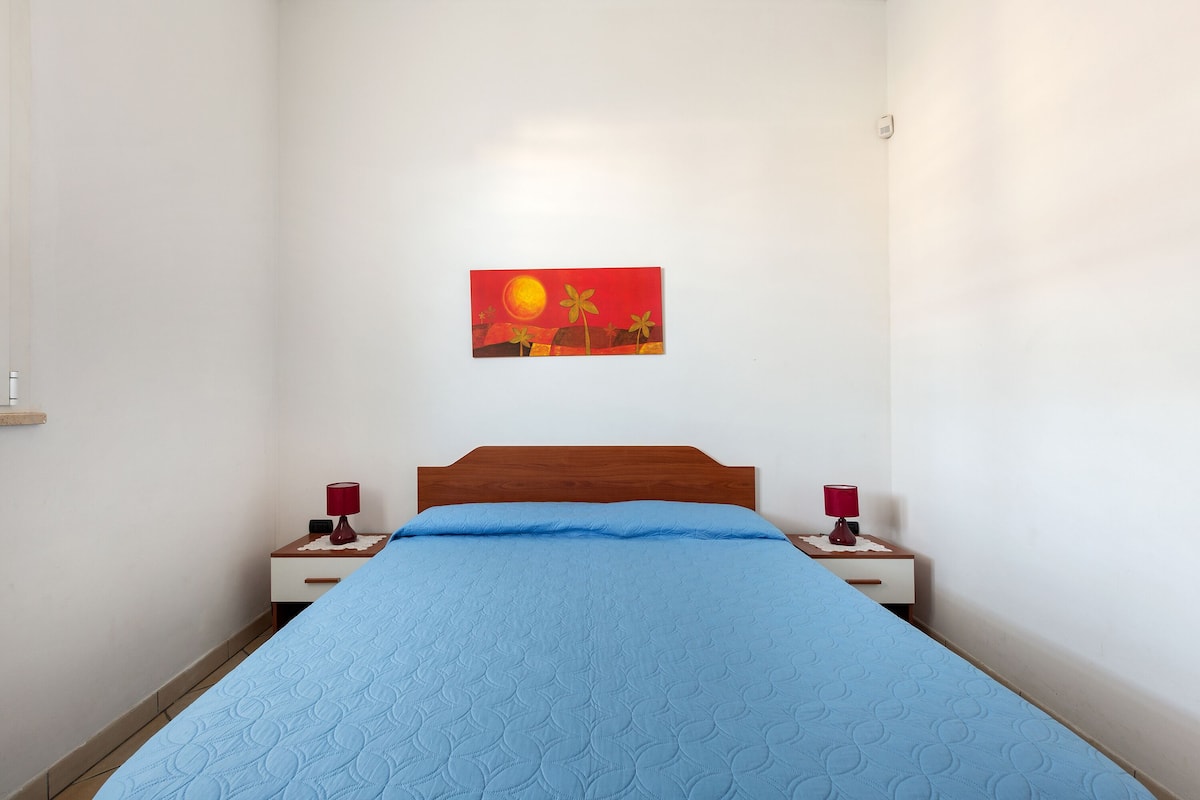 A bedroom with a bed, two bedside tables with lamps, a wall-mounted painting, and a wooden headboard.