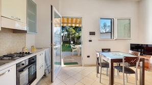 A kitchen with a stove, oven, and cabinets, a dining area with a table and chairs, and a view of a garden through an open door. - Villa Tulipano (Nardò)
