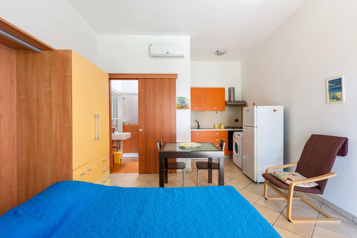 A compact room with a bed, a dining table, a kitchen area, and a chair.