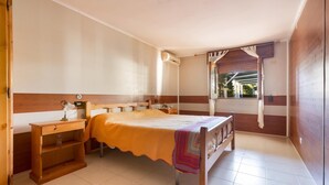 A bedroom with a wooden bed, a nightstand, a shelf, and a window with a view of greenery. - Villetta Spiaggia Bella (Lecce)