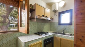 A compact kitchen with a gas stove, oven, and sink. There is a window with a view of trees and a white ceiling fan. - Villetta Spiaggia Bella (Lecce)