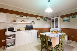A kitchen with white cabinets, a microwave, a sink, and a dining table with chairs. There are shelves above the counter and a clock on the wall.