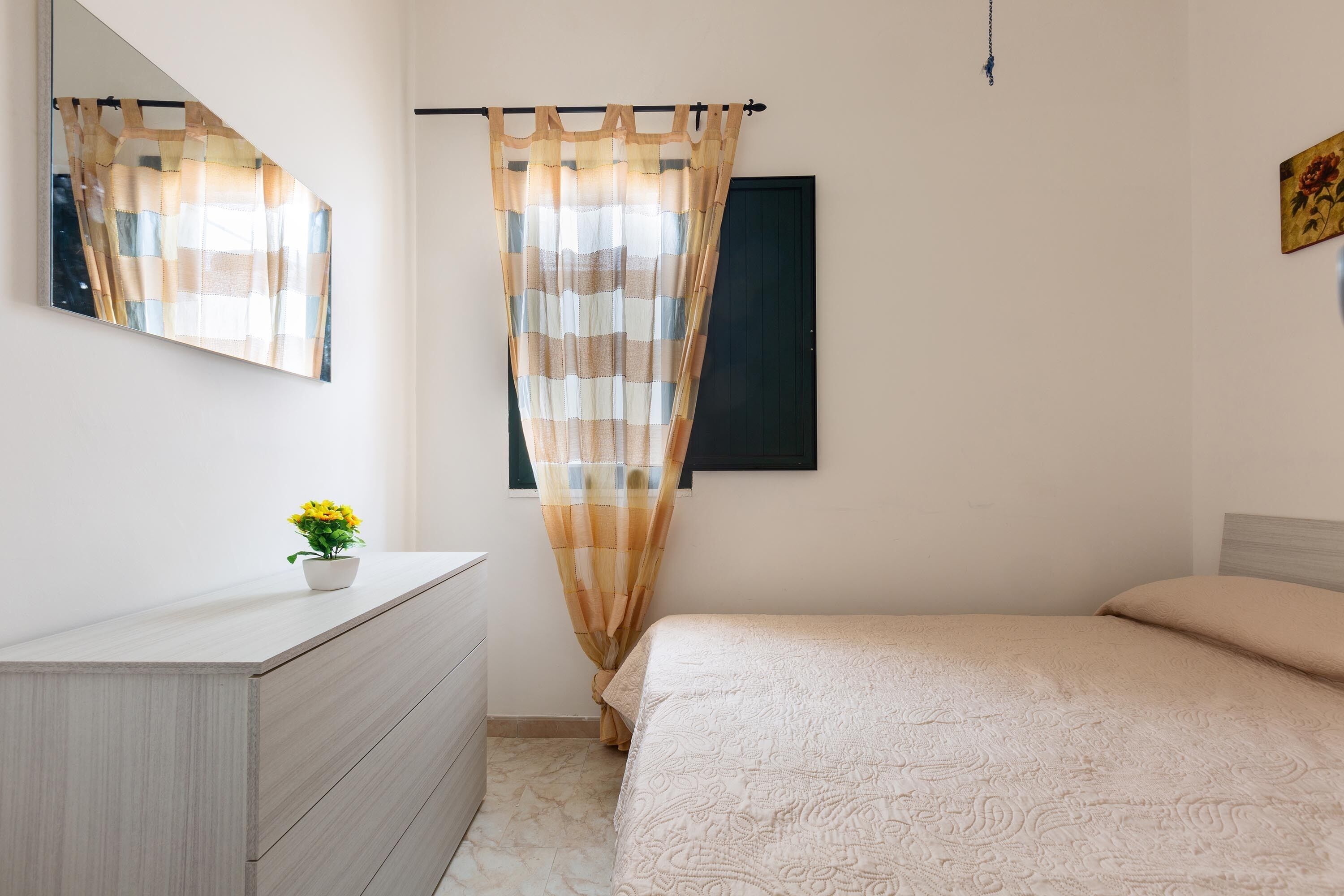 A bedroom with a bed, a dresser, a mirror, a window with curtains, and a small plant on the dresser.