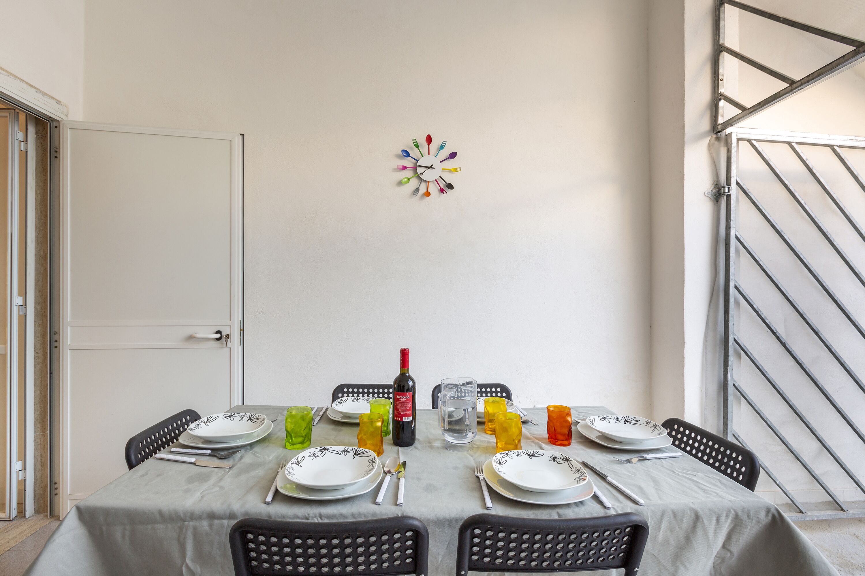 A dining table set with plates, cutlery, and colorful glasses, with a wine bottle and a clock on the wall.