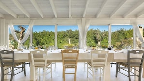 A dining area with a white table set for six, surrounded by white and dark wooden chairs, large windows with white curtains, and a view of greenery outside. - 1738 Tenuta degli Ulivi by Perle di Puglia (Melendugno)