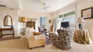 Een woonkamer met een bank, fauteuil en een televisie. Er is een plafondventilator, een tafel met een lamp en een raam met uitzicht.