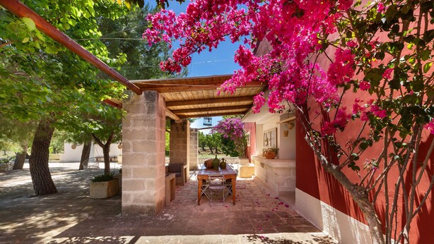 A covered patio area with a table and chairs, surrounded by trees and a red wall. - Villa Thea Charming House (Carovigno)
