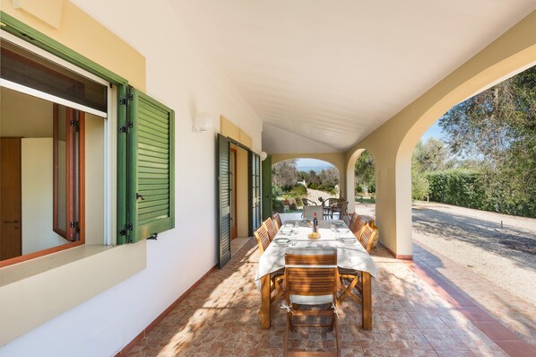 A covered patio with a table set for dining, wooden chairs, and a view of the garden.