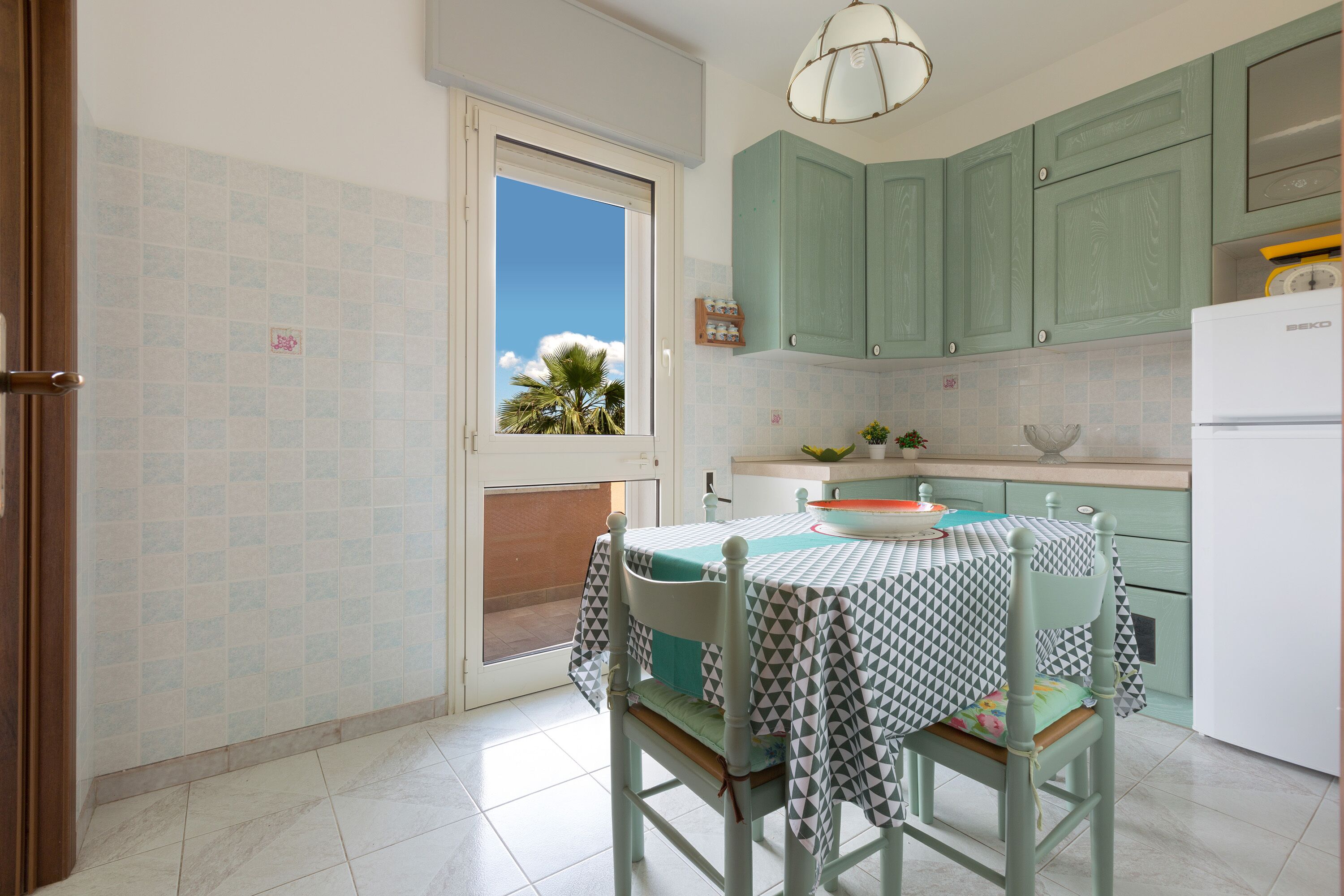 A kitchen with a table set for two, a window with a view of palm trees, and green cabinets.