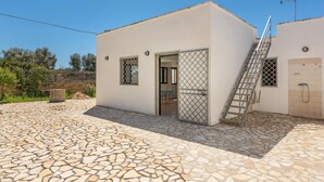 A small, single-story building with a stone-tiled patio, a door with a metal gate, and a staircase leading to an upper level.