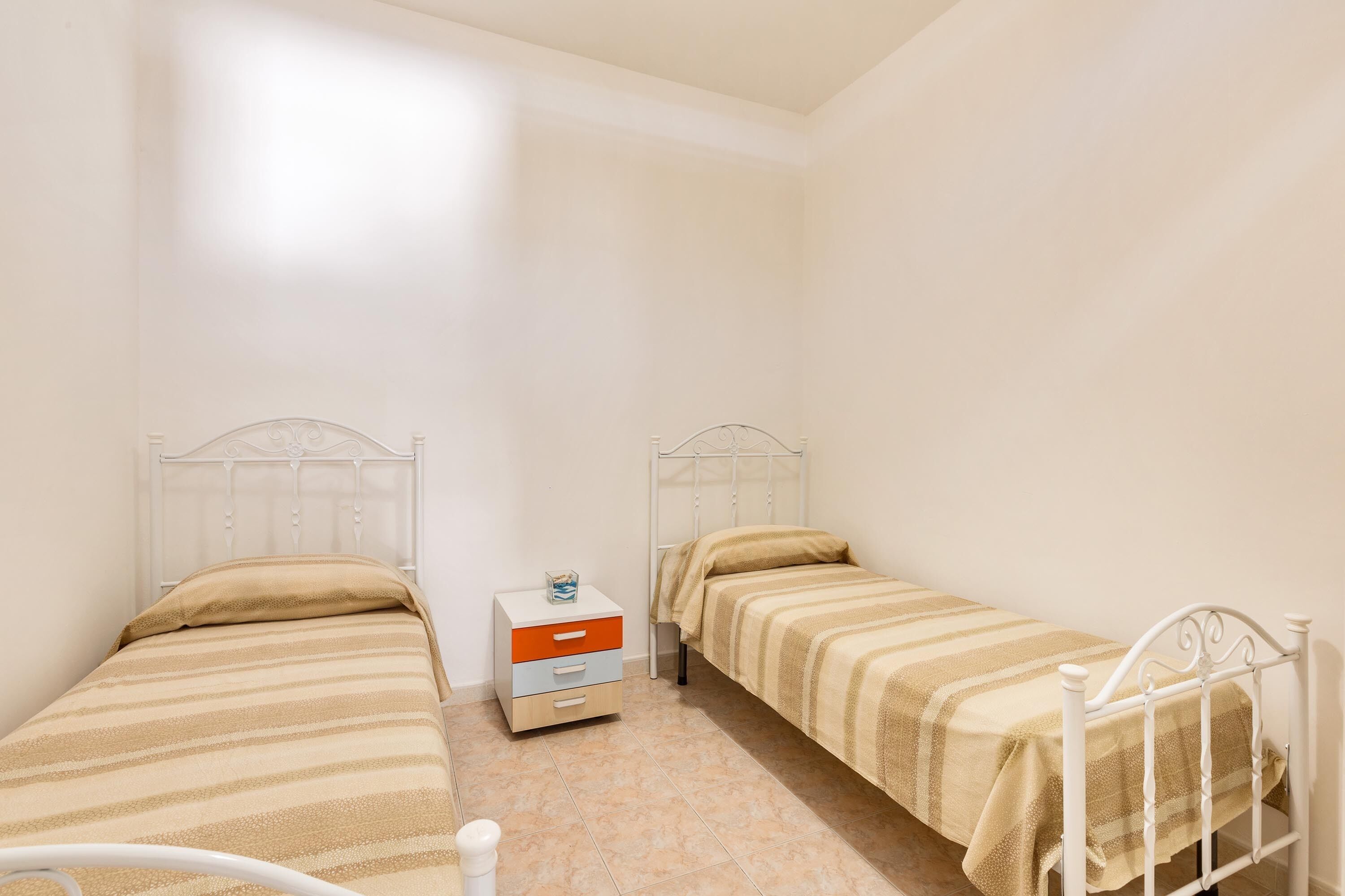 Une petite chambre propre, avec deux lits simples, une table de chevet et une commode.
