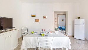 A dining area with a white table, chairs, and a TV on a cabinet. There are framed pictures on the wall and a refrigerator in the background. - Casa Medusa (Torricella)