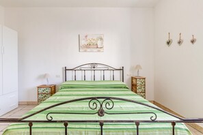 A bedroom with a metal bed frame, green striped bedding, a nightstand with a lamp, a wooden cabinet, and a wall-mounted picture. - Casa Medusa (Torricella)