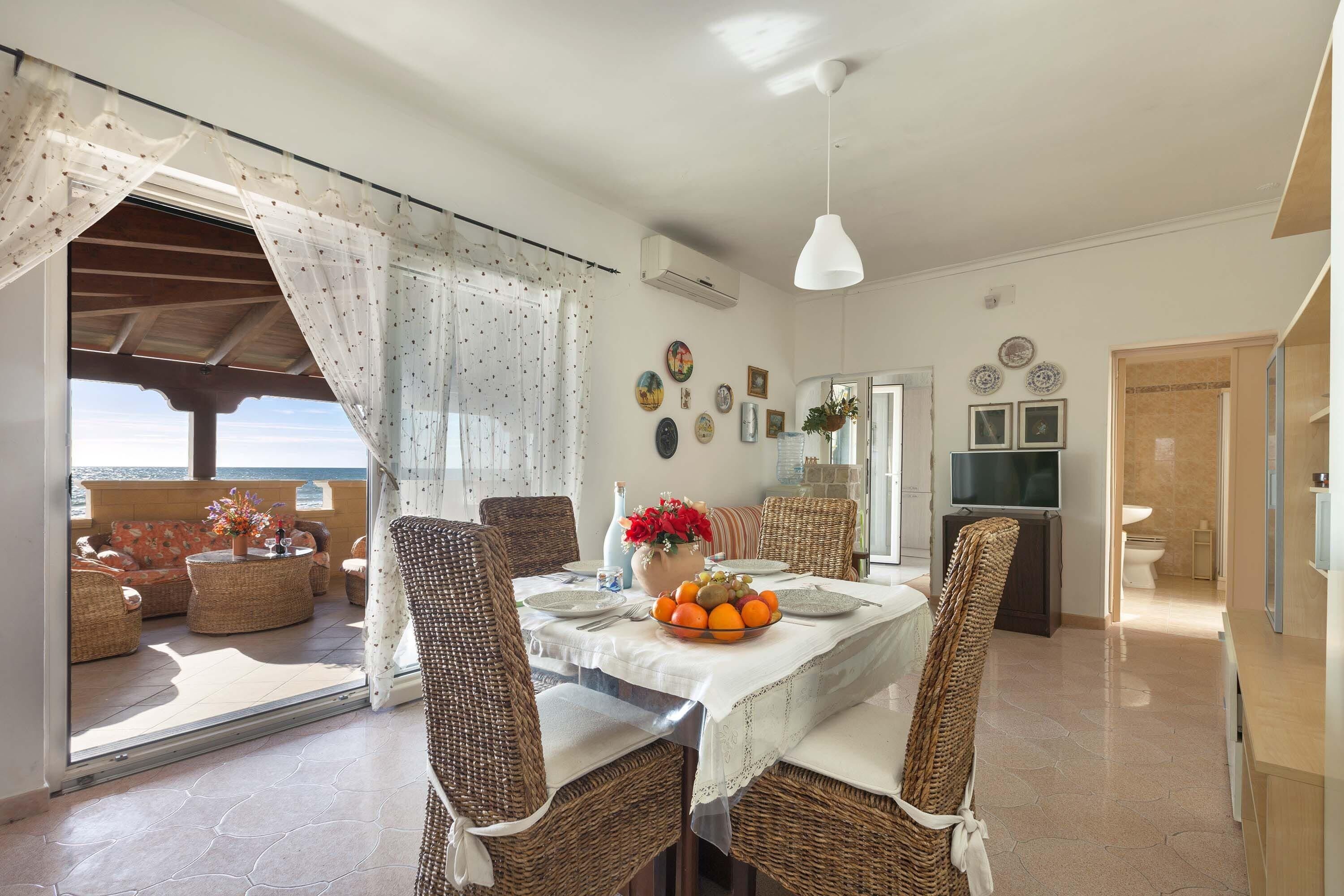 Un comedor con mobiliario de mimbre, una mesa dispuesta con platos, un frutero y un florero. Se observa el mar a través de la puerta de cristal.