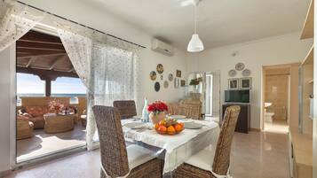 Un comedor con mobiliario de mimbre, una mesa dispuesta con platos, un frutero y un florero. Se observa el mar a través de la puerta de cristal.
